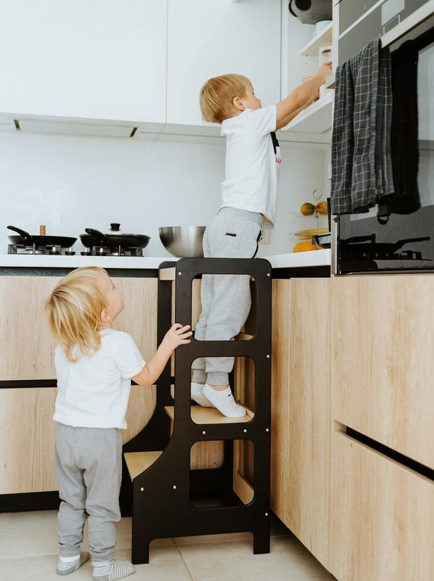 Black learning tower with children reaching for cupboard in kitchen